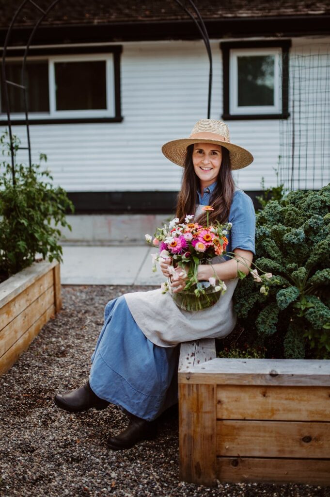 Woman in denim dress and straw hat holding pink flowers in a home garden.